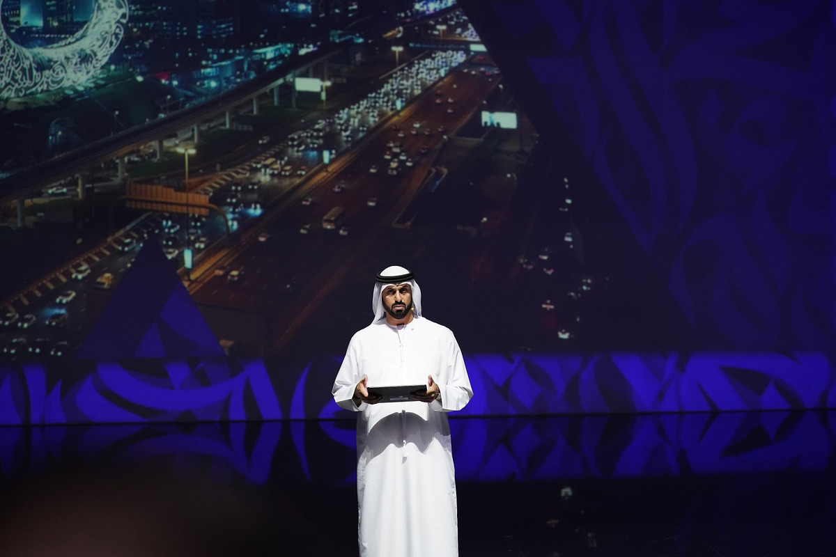 Man in Emirati attire holding a tablet on stage.