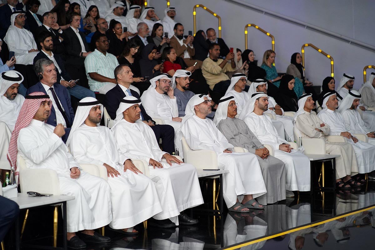 Men in thawbs sit at a formal event in a modern hall.