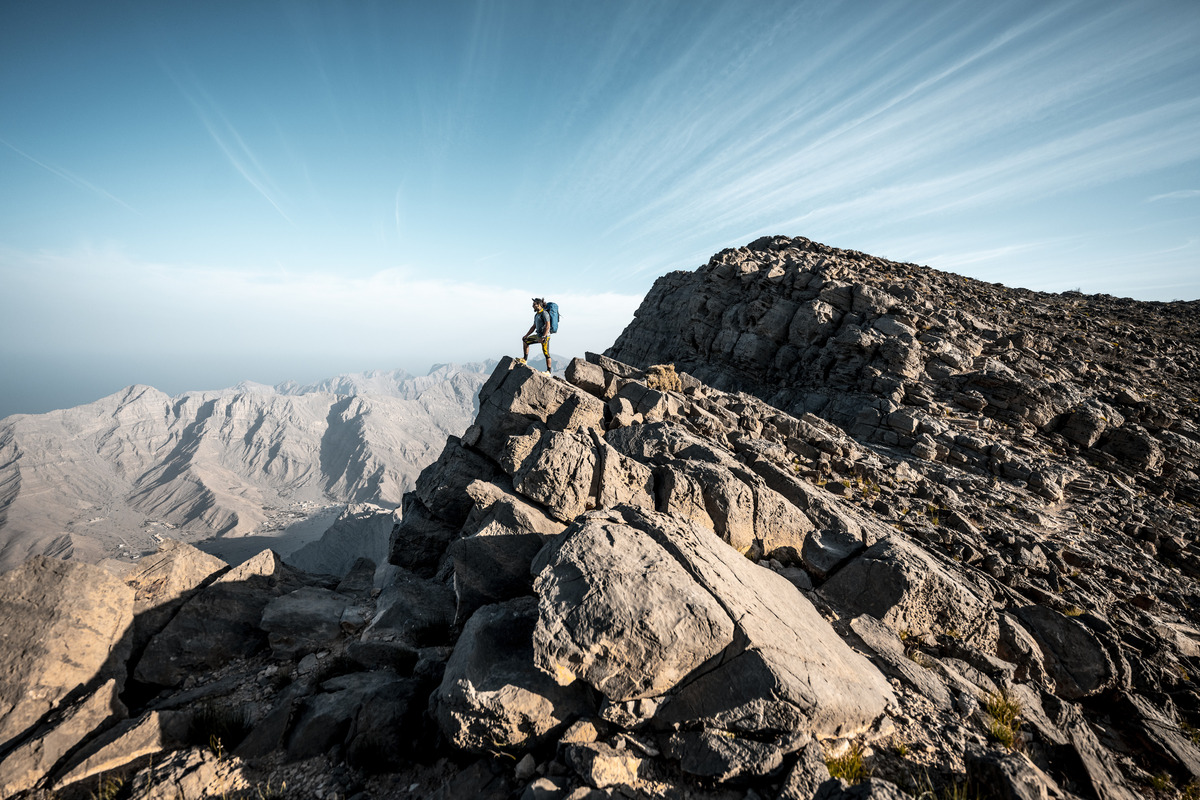 Hiker atop rocky peak, overlooking mountains.