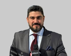 Man in a suit speaking into a microphone with a light gray background.