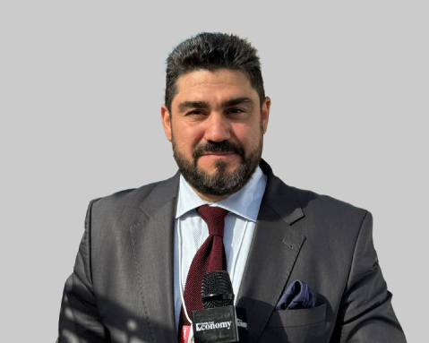 Man in a suit speaking into a microphone with a light gray background.