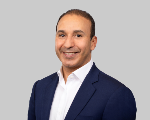 Man in navy suit smiles against gray background.