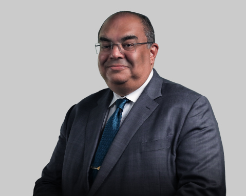 Man in a dark suit and teal tie poses against a light gray background.