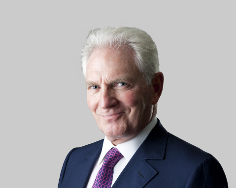 Older man in a dark blue suit and purple tie smiling against a gray background.