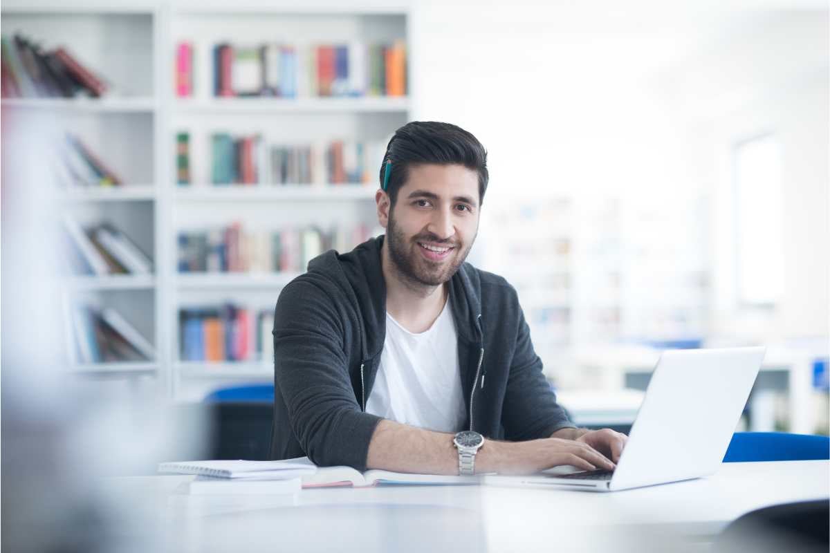 Man using a laptop in a library with bookshelves in the background.