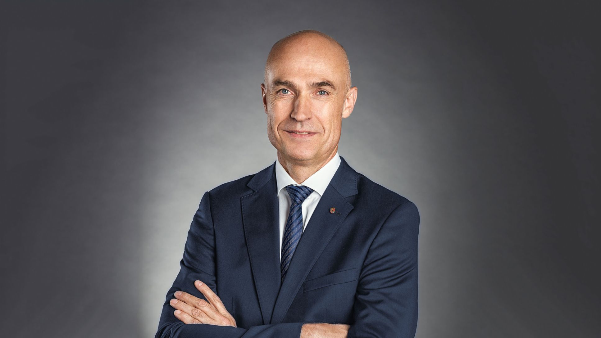 Bald man in a dark suit and tie standing with arms crossed, smiling slightly.