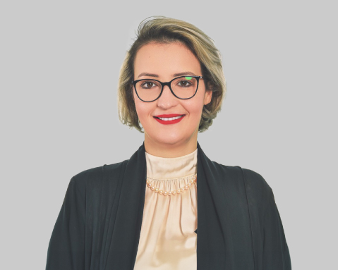 Woman in glasses and black jacket smiling against a gray background.