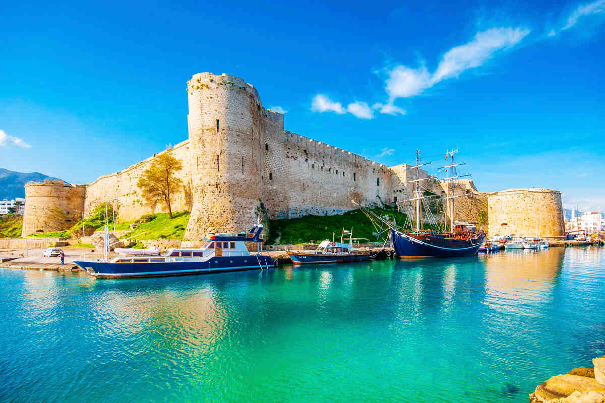 Stone castle by coast with boats in calm water under sunny sky.