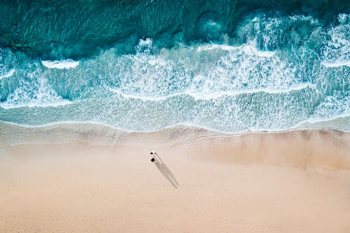 Two people walk on sandy beach with turquoise waves.