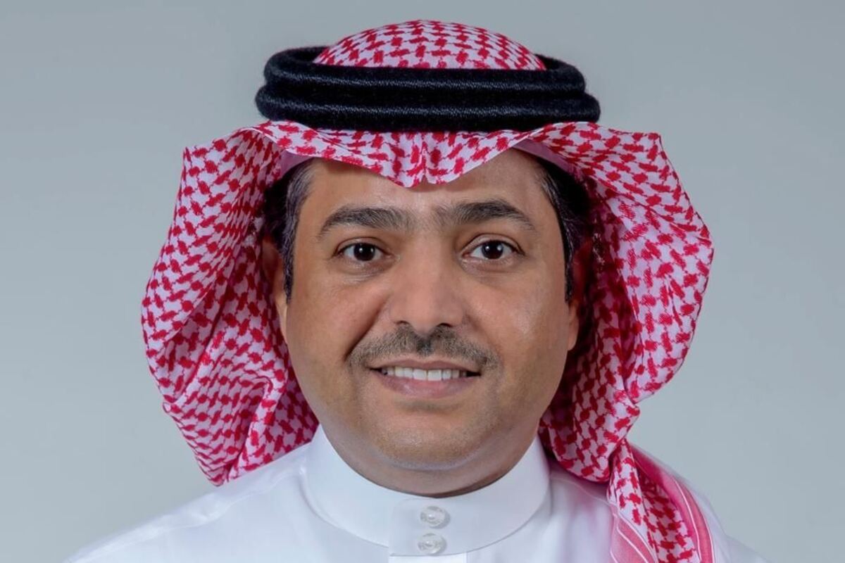 Man in patterned headdress smiling at the viewer with grey background.