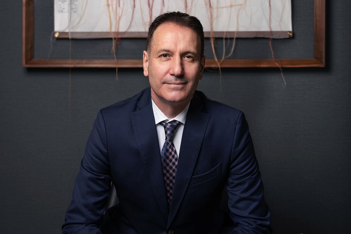 Man in suit sits before a dark gray wall with framed art.