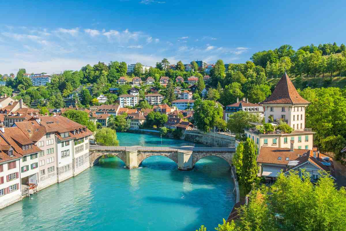 Stone bridge spans turquoise river in hillside town under blue sky.
