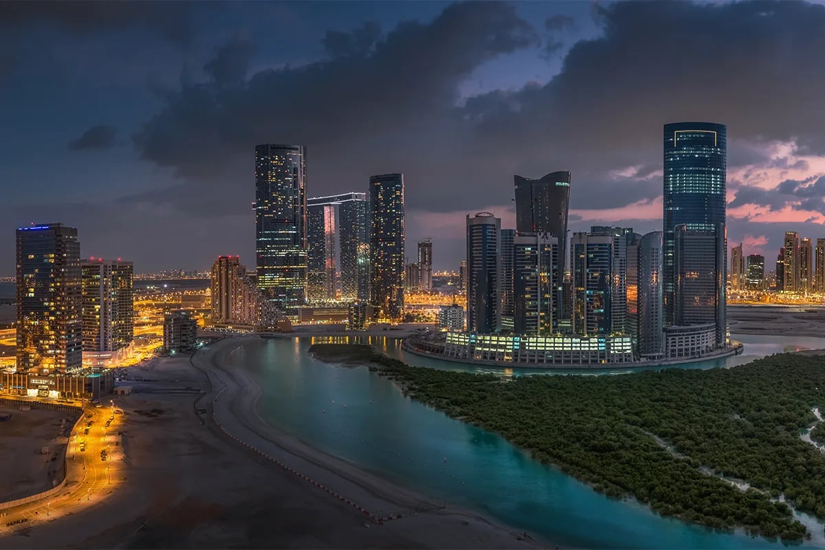 City's skyscrapers lit up at dusk beside a calm waterway.