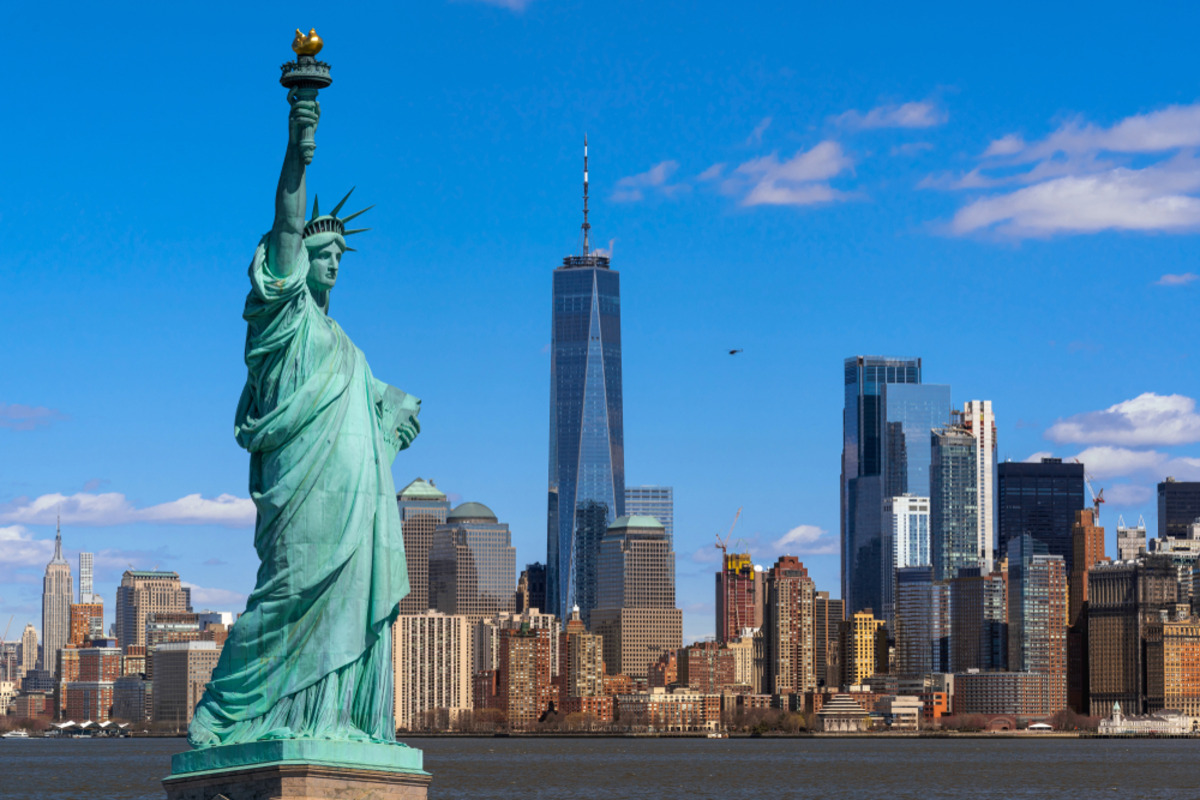 The Statue of Liberty stands on a pedestal with a cityscape under a blue sky.