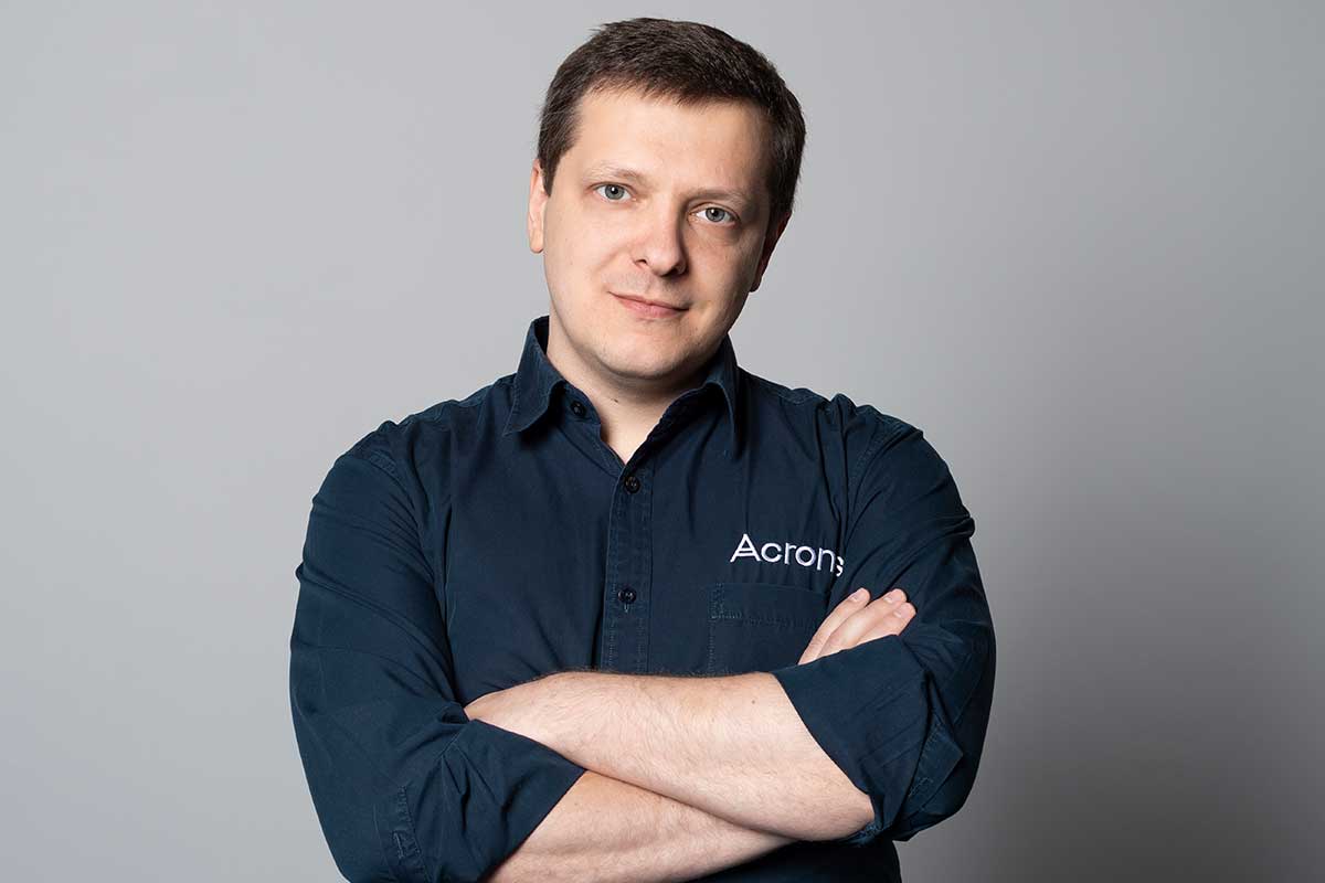 A man in a blue shirt with "Acronis" stands with arms crossed against a gray background.