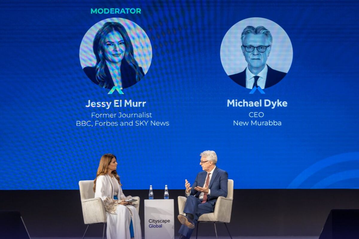 Two individuals converse onstage at a conference, with headshots displayed behind them.