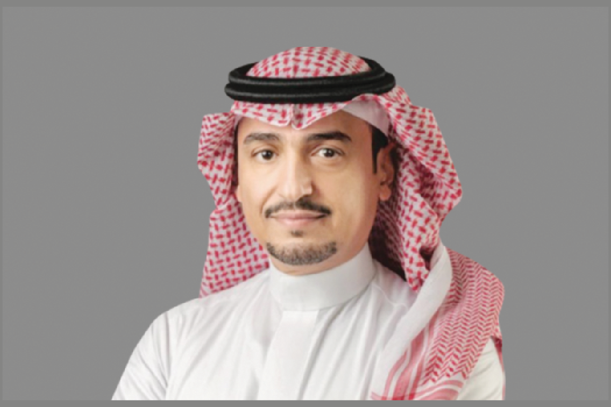 Saudi man in keffiyeh poses against gray.